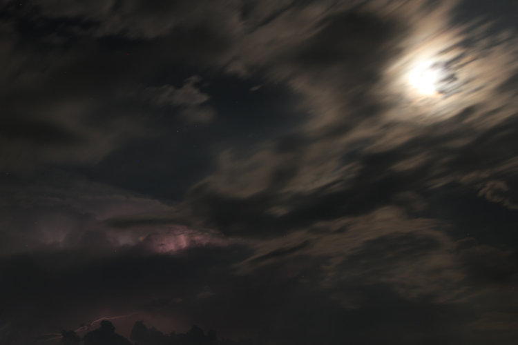 hint of lightning under distant clouds while moon peeks out from behind fast-moving clouds during time exposure at night